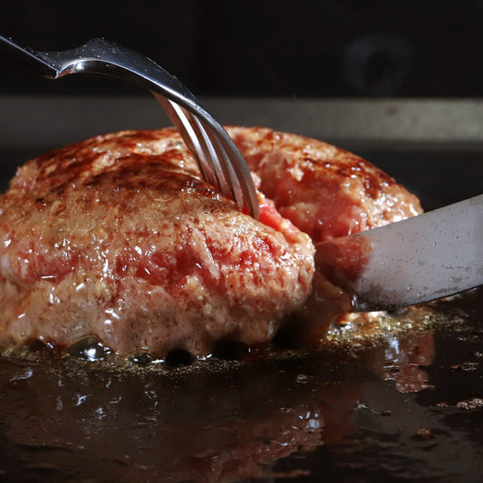 松阪牛ハンバーグを鉄板でフォークとナイフで切り開いた瞬間、溢れ出す肉汁とロゼ色の断面がクローズアップで映し出される