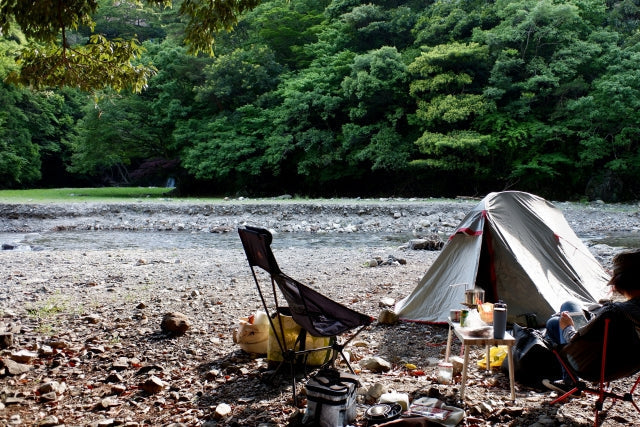 川沿いの砂利河原にテントとアウトドアチェアを設置し、一人でくつろぐソロキャンプの様子。緑豊かな山を背景に、のんびりとした自然の中での食事時間が伝わってくる