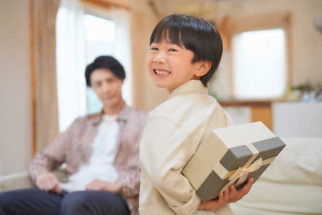 男の子がグレーのリボン付きギフトボックスを後ろ手に隠しながらうれしそうに振り返り、奥でお父さんが微笑む
