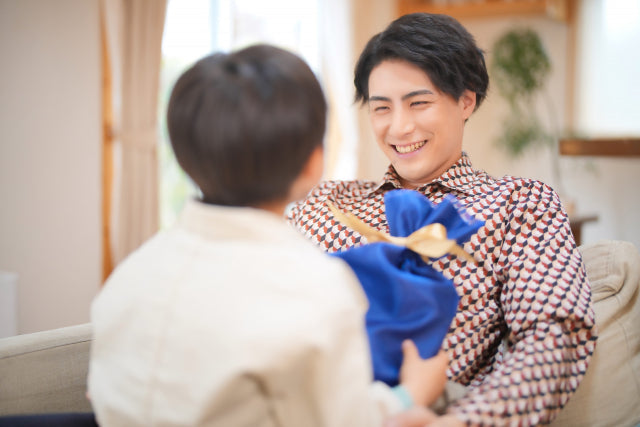 子どもがソファに座った父親に青い風呂敷包みのプレゼントを手渡し、父親が笑顔で受け取っている父の日のシーン