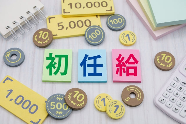 初任給の文字が入ったカラフルなタイルを中心に、硬貨や紙幣のミニチュアと電卓が周囲に散りばめられている
