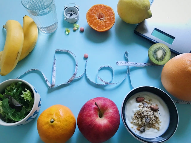 メジャーで「Diet」の文字を形作り、バナナやオレンジ・キウイ・リンゴ・レモンなどの果物とグラノーラヨーグルト・サラダを水色の背景に並べたダイエット食材の俯瞰