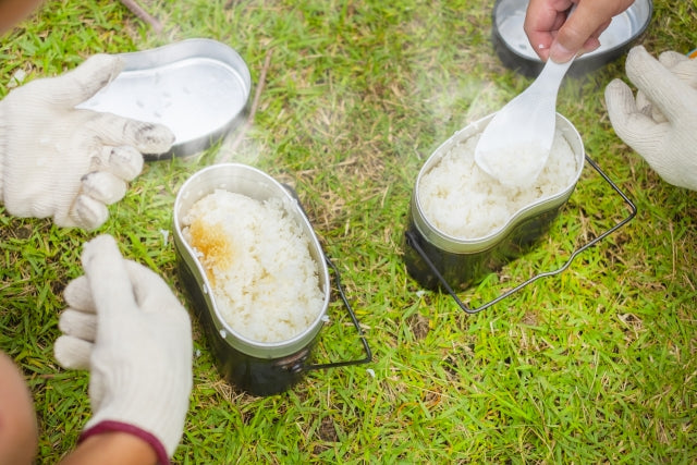 キャンプ場の芝生の上で飯盒炊爨した炊きたてご飯を軍手をした手でスプーンによそっている様子