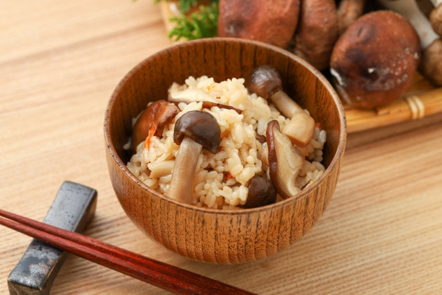しめじや人参が入った炊き込みご飯が木製の茶碗に盛られ、背景には椎茸が並ぶ