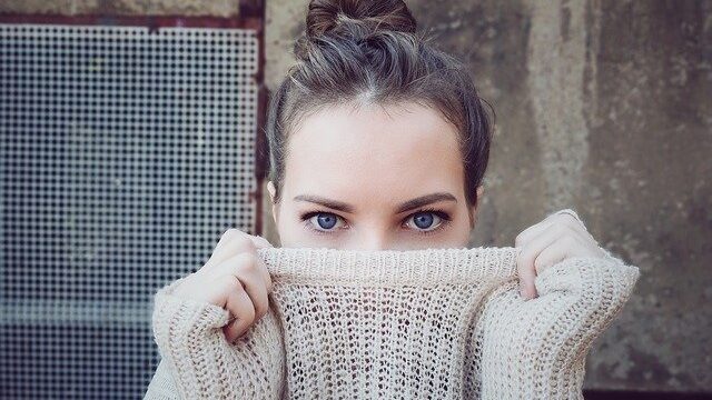ニット素材の襟元を両手で引き上げて顔の下半分を隠す青い目の女性が、カメラを見つめている