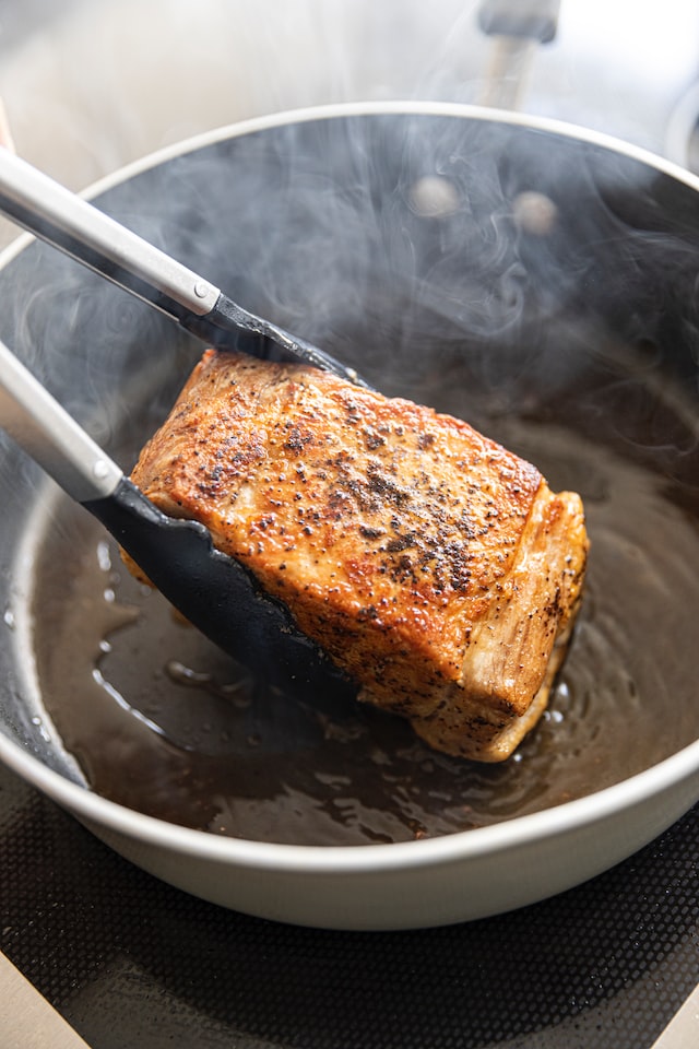 トングで持ち上げられた焼きたてのステーキが、煙の立ち上るフライパンの上で美しい焦げ目をつけて仕上がっていく様子