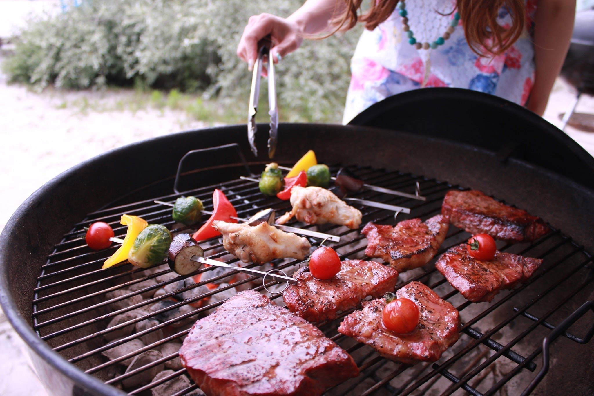 丸型バーベキューグリルの上で肉や野菜串を焼く女性がトングで食材を扱っている自宅BBQの様子