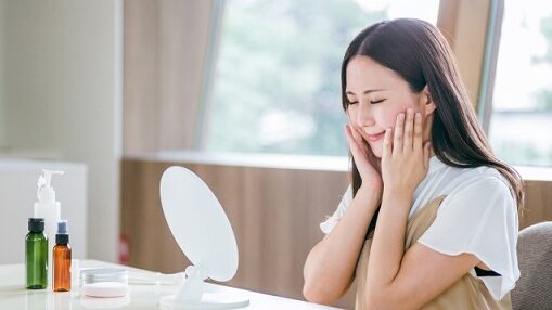 鏡の前で両手を頬に当て、なめらかな肌をうれしそうに確認する若い女性。テーブルにはスキンケア用品が並んでいる