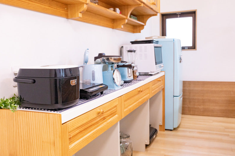 木製造作棚のキッチンカウンターに黒い炊飯器とブルーのケトルや調理家電が並び、奥にレトロデザインの水色冷蔵庫が見える