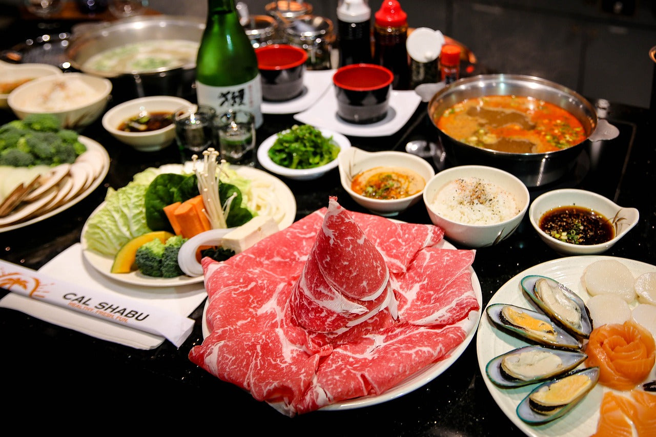 黒いテーブルに並んだしゃぶしゃぶ食材の全景。中央の白い皿に霜降り和牛の薄切り肉が盛り付けられ、奥に赤いスープと白いスープの2種の鍋、野菜の盛り合わせ、ムール貝と魚介の皿が広がる