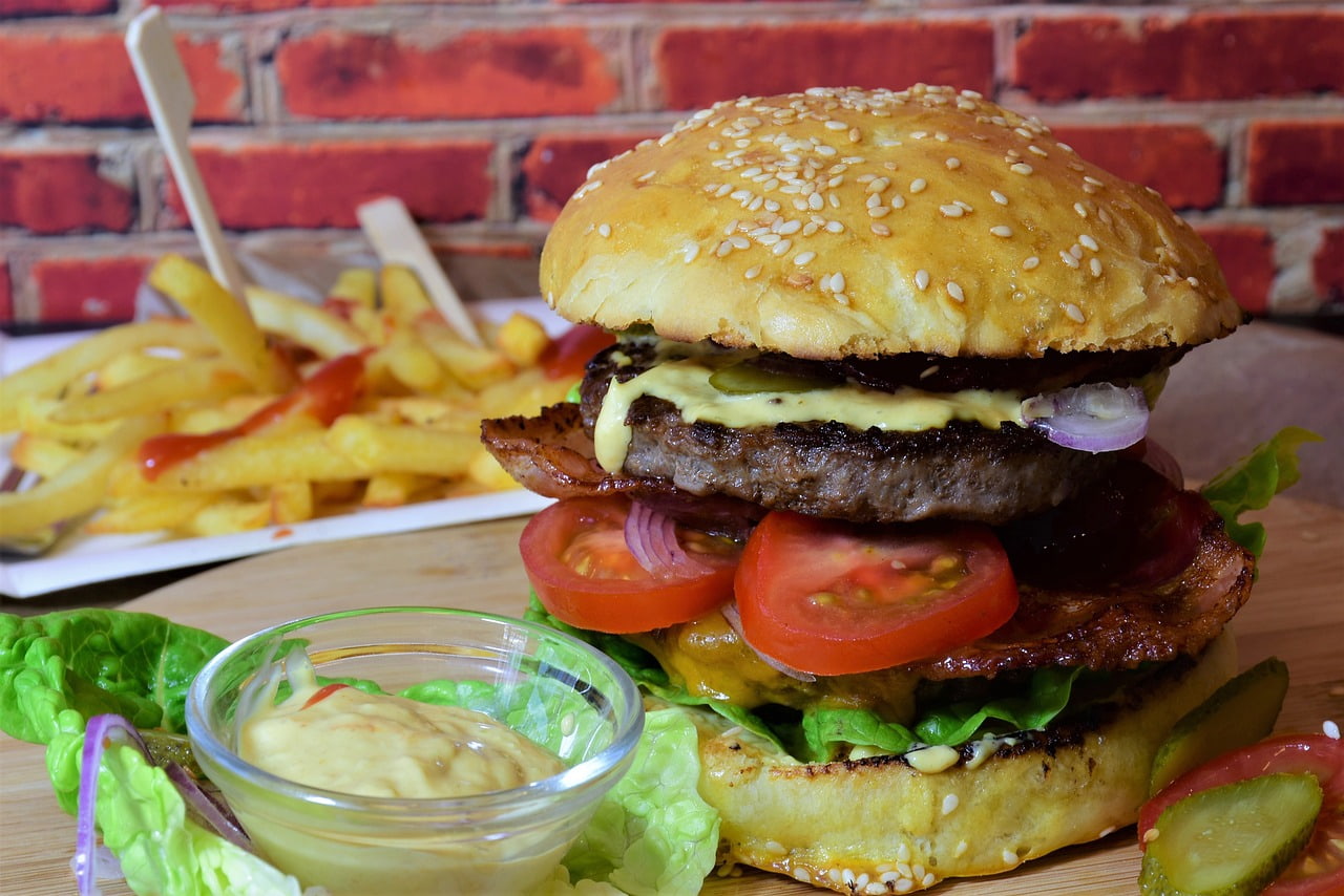 ゴマ入りバンズに肉厚なビーフパティとトマト・レタス・オニオンを重ねた食べ応えのあるハンバーガーが木製トレーに盛られ、脇にマスタードソースとフライドポテトが添えられている