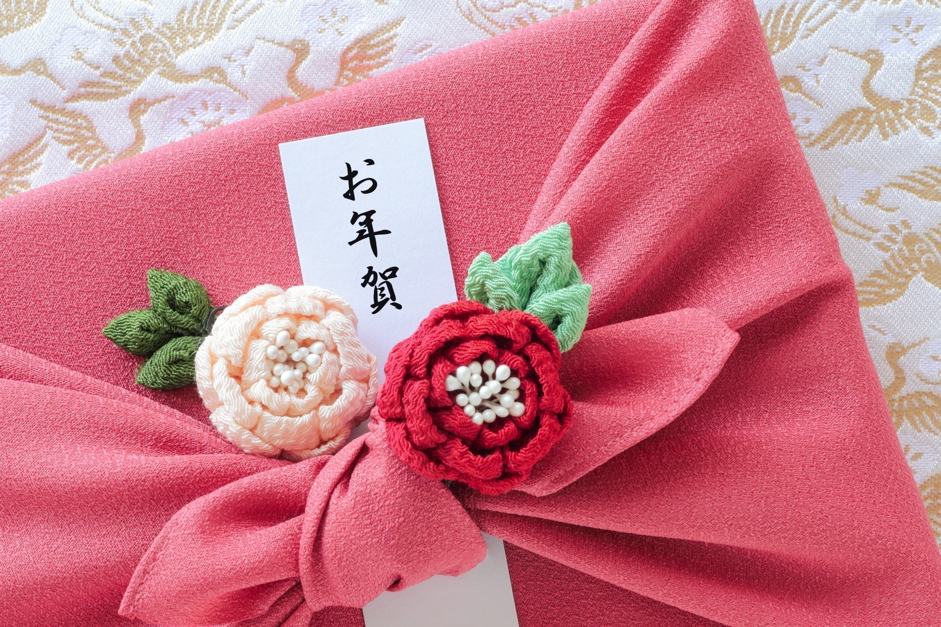 ピンクの風呂敷に包まれたお年賀ギフトと、手作りの刺繍花飾りが添えられたお年賀の熨斗カード