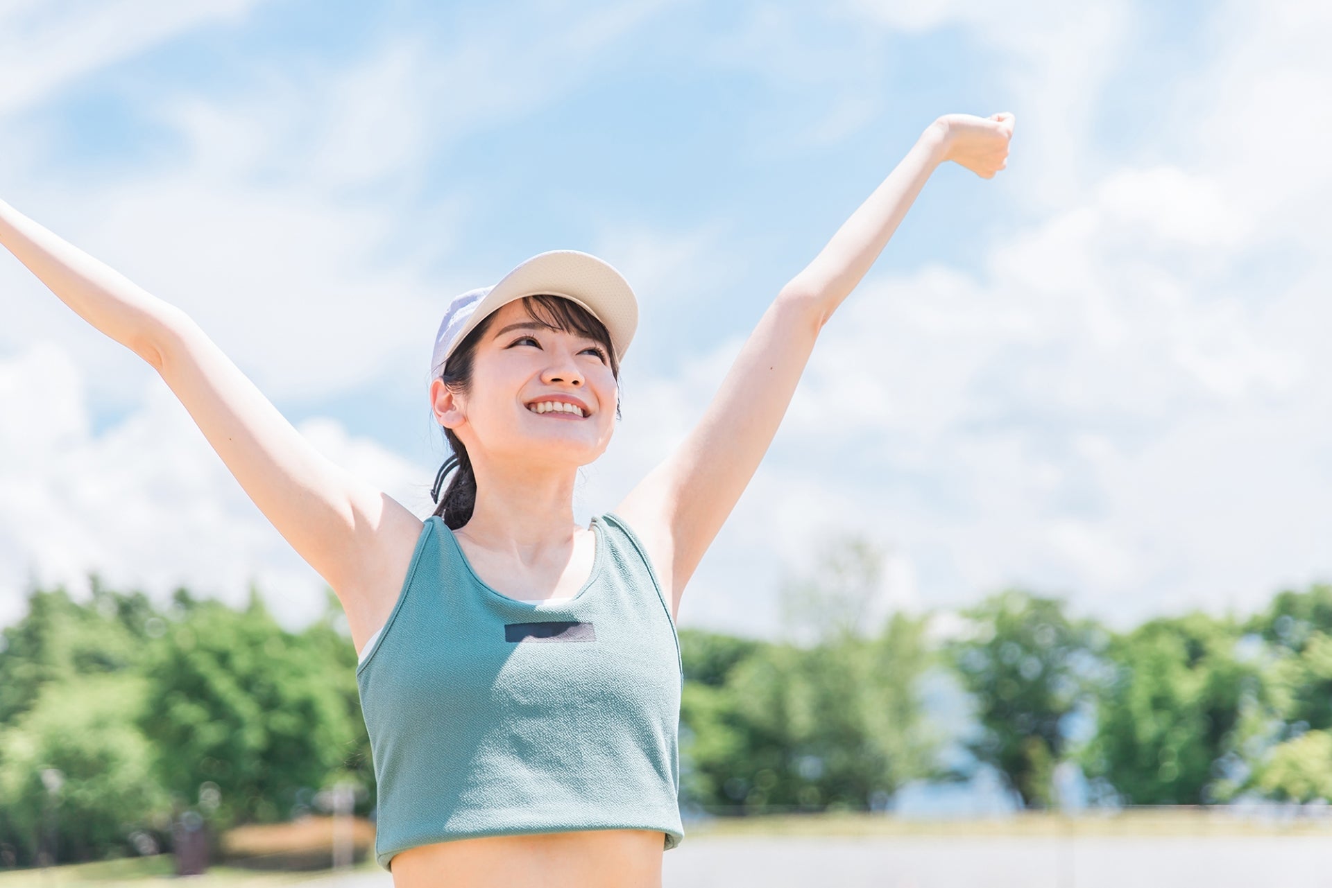 青空の下で両腕を大きく広げて笑顔を見せるスポーツウェア姿の女性が健康的な爽やかさを表現している