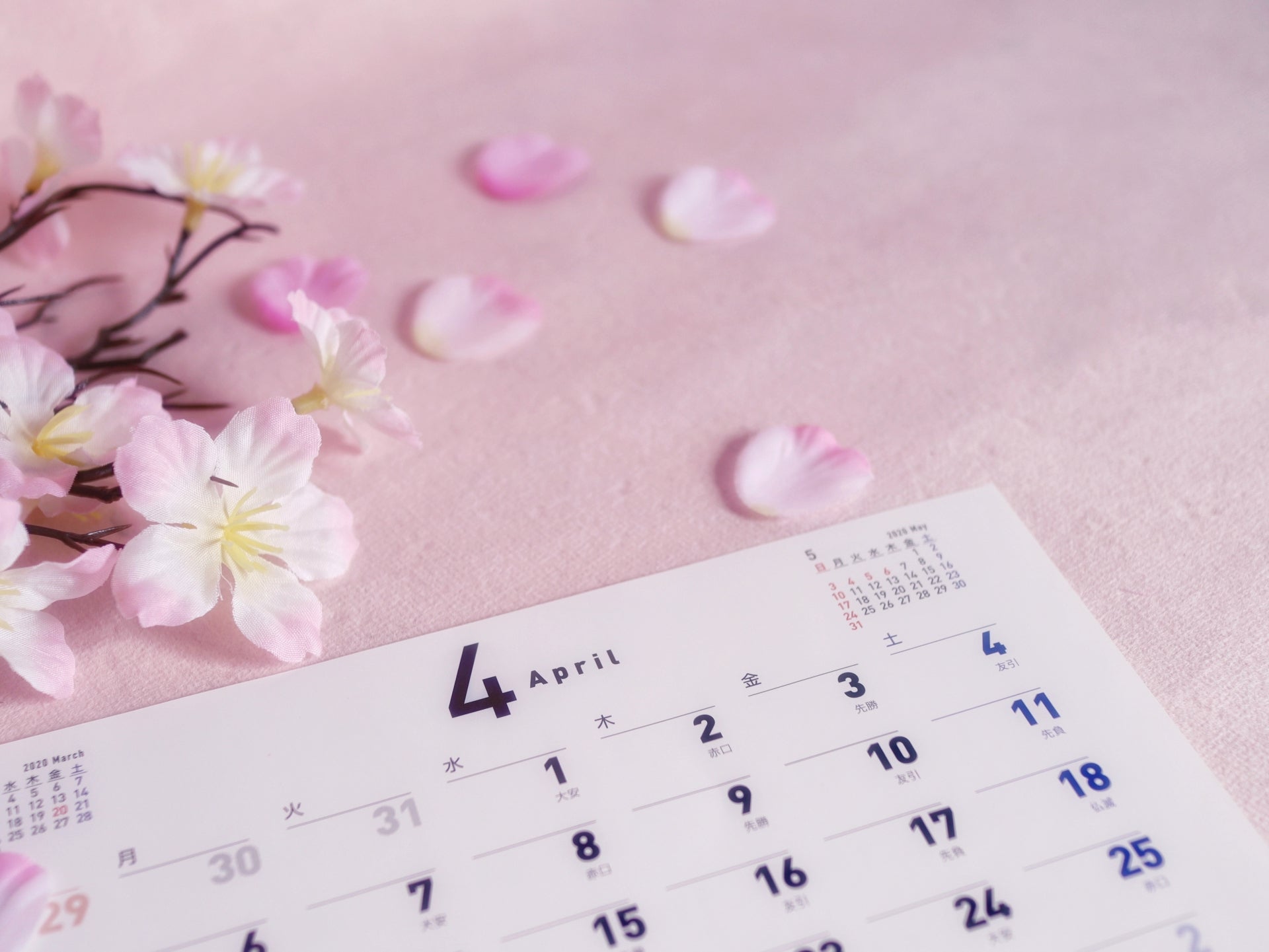 桜の花びらが散る淡いピンクの背景に4月のカレンダーが置かれ、春の節目シーズンを表している