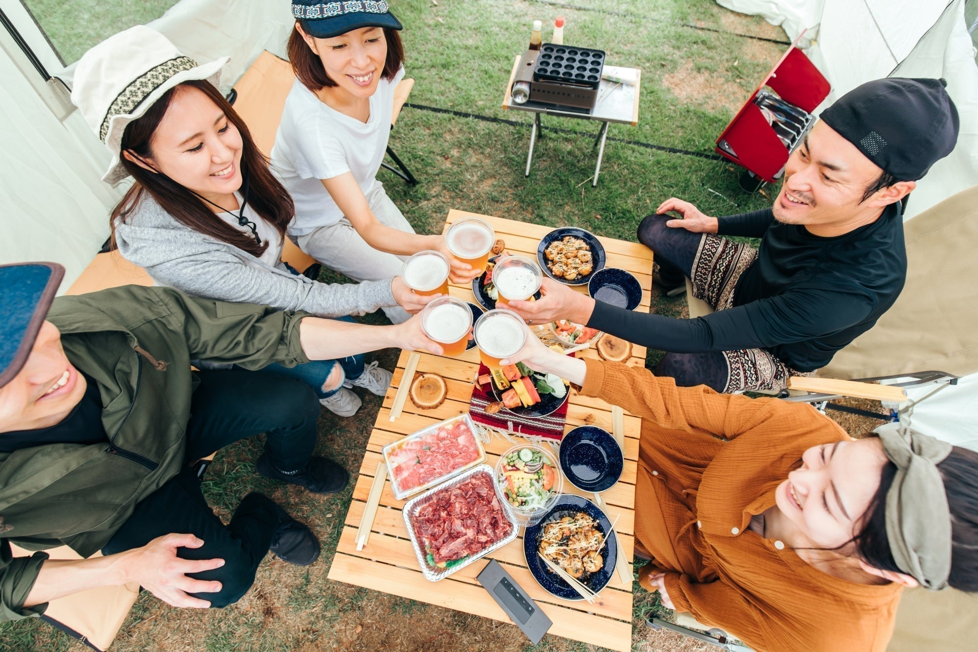 キャンプテントの下で5人の男女がビールを持ち寄って乾杯し、テーブルには生の和牛スライスや焼き物料理が並ぶアウトドア焼き肉パーティのシーン