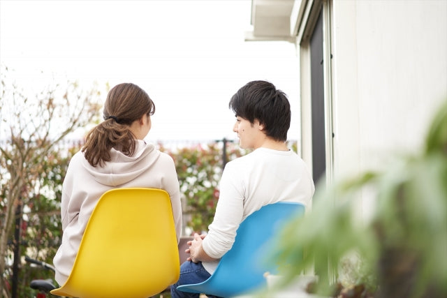 屋外のテラスで黄色と青色の椅子に腰掛け、向かい合って会話を楽しむカップルがおうちデートのひとときを過ごしている