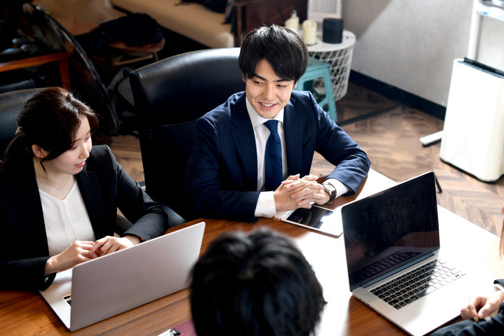 スーツを着た男性と女性がオフィスのテーブルを囲み、ノートPCやタブレットを前にしながら打ち合わせをしている場面で、中央の男性が笑顔で手を組んでいる