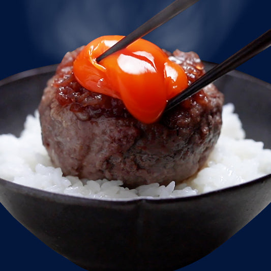 黒い器に盛った白米の上に丸いハンバーグを置き、箸で卵黄を乗せる瞬間を湯気とともに接写で捉えた