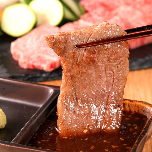焼き上がった和牛の薄切り肉を箸でつまみ、醤油ベースのたれに浸している。艶やかな焼き色と滴る肉汁が際立ち、背景に生の霜降り肉と野菜が並ぶ