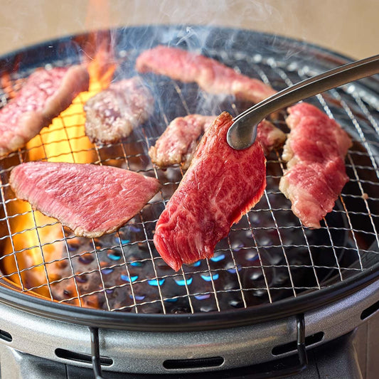 鳥取和牛のモモ焼肉が七輪の網の上で炭火と炎を受けながら焼かれ、赤身と霜降りのバランスが映える肉厚なスライスをトングでつまんでいる場面