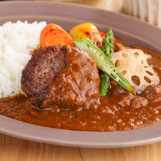 飛騨牛ハンバーグがカレーソースに浸り、アスパラや蓮根などの焼き野菜を添えた贅沢な一皿のアップ
