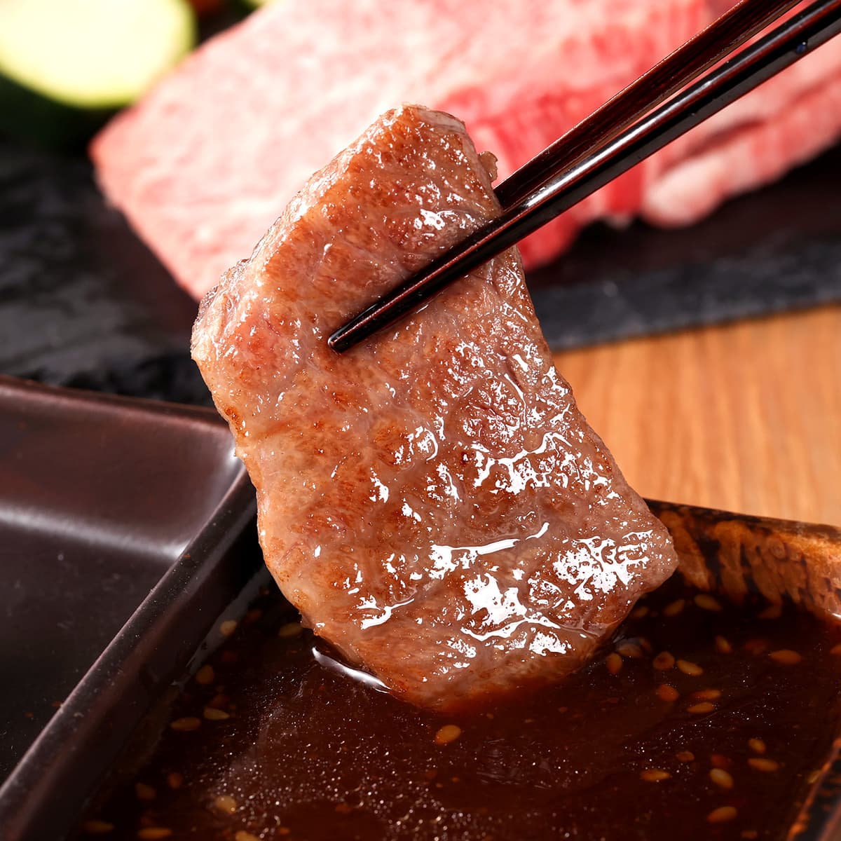 箸でつまみ上げた山形牛カルビの焼き上がり。照りのある褐色の表面からジューシーな脂がしたたり落ち、奥には生の霜降り肉が見える