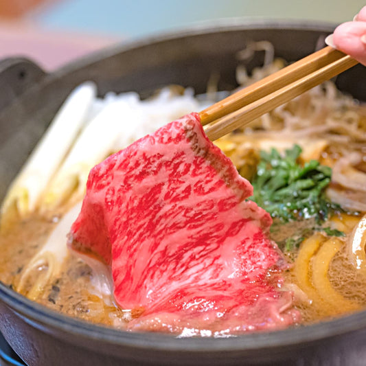 すき焼き鍋の割り下に赤身スライスと白ネギ・しらたきが煮えており、箸で黒毛和牛の薄切り肉を持ち上げているところ